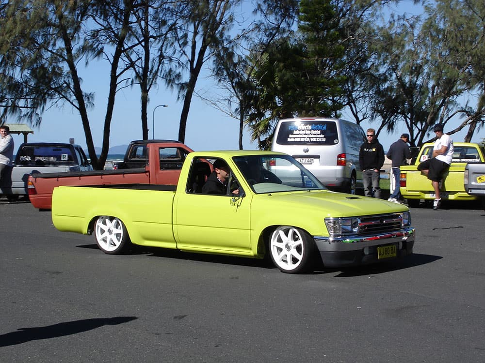 Bright yellow lowered Toyota Hilux with white chrome wheels at Half Way Havoc waterfront gathering