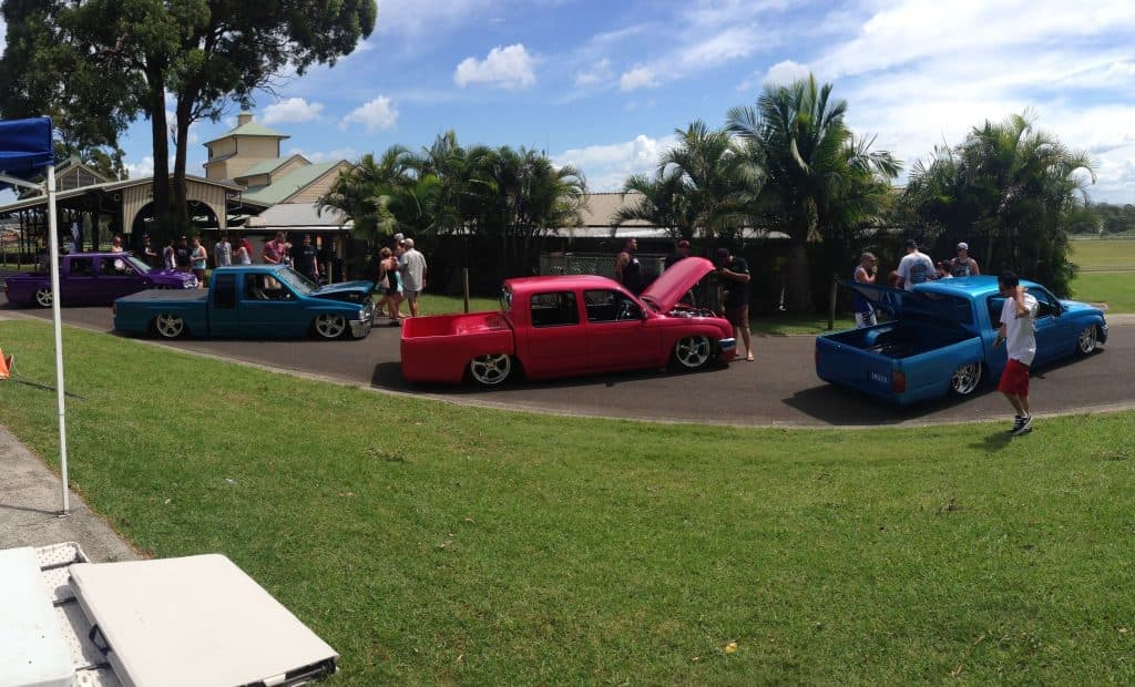 Lineup of colorful slammed Holden Rodeo and Toyota Hilux dual cabs in blue pink and teal on grass with palm trees at Port Macquarie raceway